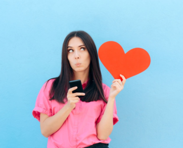 A young lady holding smartphone and a heart in her hands and thinking about dating apps