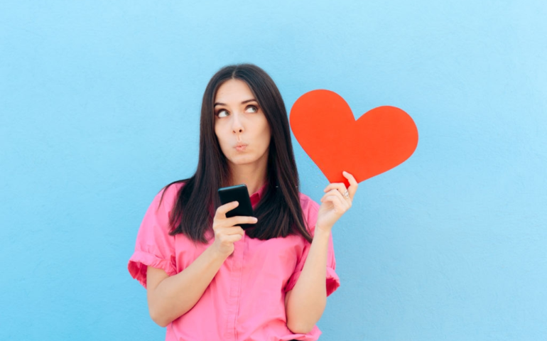 A young lady holding smartphone and a heart in her hands and thinking about dating apps
